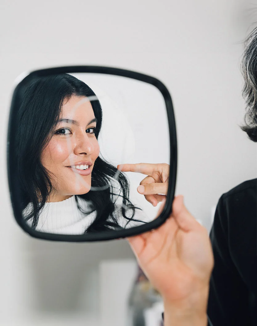 A woman with dark hair and light skin smiles while looking at her reflection in a black-framed handheld mirror. A professional, whose hand is visible, gestures near her face, possibly discussing her appearance. The background is light and blurred, suggesting a clinical or aesthetic setting with soft, even lighting. - Dermal Fillers in King of Russia, PA