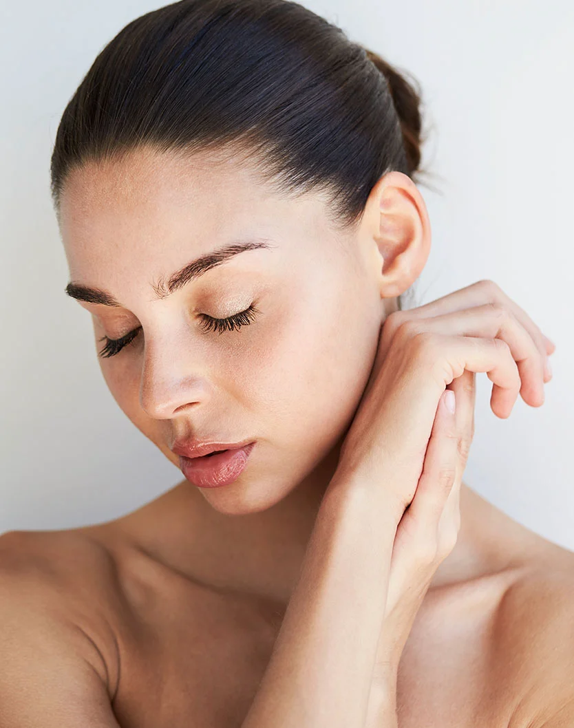 Close-up of a serene woman with smooth, light skin, closed eyes, and natural pinkish-nude lips. Her dark hair is pulled back in a bun, and her hands rest gently on her neck and shoulder. The plain light gray or white background and soft, even lighting highlight her features. - Hydrafacial in King of Russia, PA
