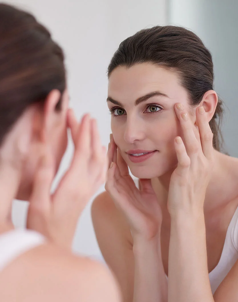 A woman examines her face in a mirror, gently touching her skin. She has dark brown or black hair pulled back and wears a light-colored tank top. The lighting is soft and natural, with a neutral-toned background, likely in a bathroom, focusing on her self-care routine. - Lutronic Lacemd in King of Russia, PA