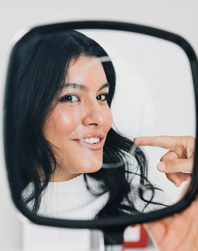 A young woman with dark hair, wearing a light cream turtleneck, smiles in a handheld mirror. Her skin has a natural glow, and she touches her cheek with one hand. The dark gray mirror reflects her friendly expression against a simple, neutral background with soft lighting. - Microdermabrasion in King of Russia, PA