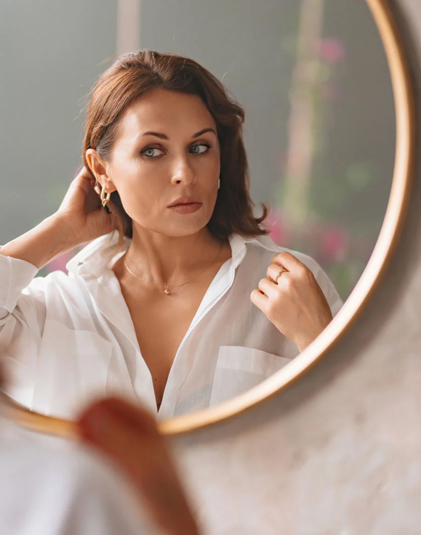 A middle-aged woman with shoulder-length brown hair looks at herself in a round gold-framed mirror. She wears a loose white shirt, a gold pendant necklace, and a gold ring. The soft, warm lighting highlights her serious expression, with a muted background and a second person's hand subtly visible. - Minoxidil in King of Russia, PA