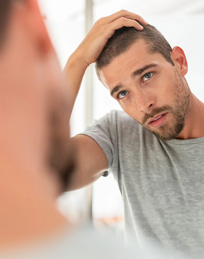 A man with a short buzzed haircut and light-medium beard looks at his reflection in a mirror with a thoughtful or concerned expression. He has light blue eyes and wears a gray, short-sleeved t-shirt. His hand is on his head, likely adjusting his hair. A blurred reflection of his head and upper torso is visible in the mirror, with a light, neutral-colored background out of focus. - Nutrofol in King of Russia, PA