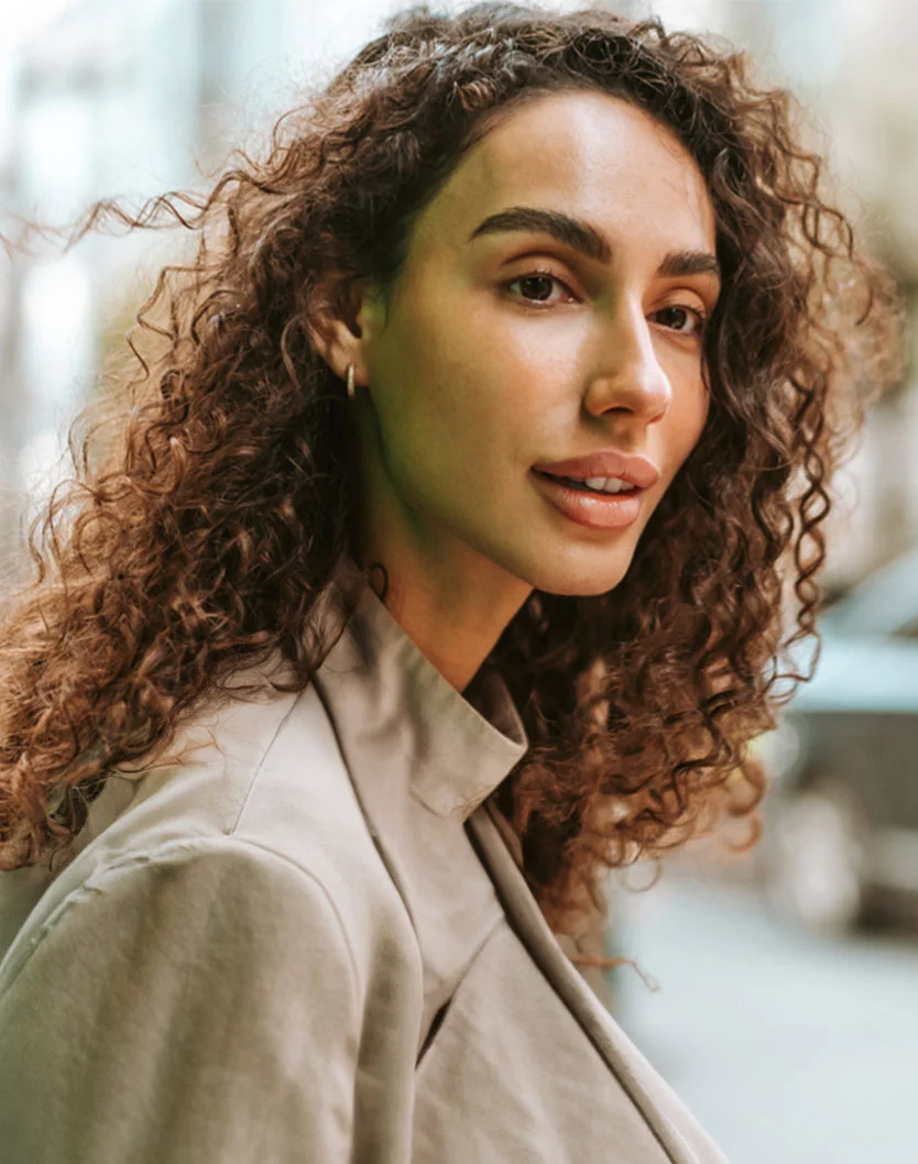 A light-skinned woman with curly, dark brown hair smiles gently, her expression pleasant and approachable. She wears a light beige or taupe jacket with a collared neckline, and a small hoop earring is visible on her earlobe. The background, blurred with urban structures, suggests a city setting. Natural lighting highlights her features, creating soft shadows. - PDO Threads Nova in King of Russia, PA