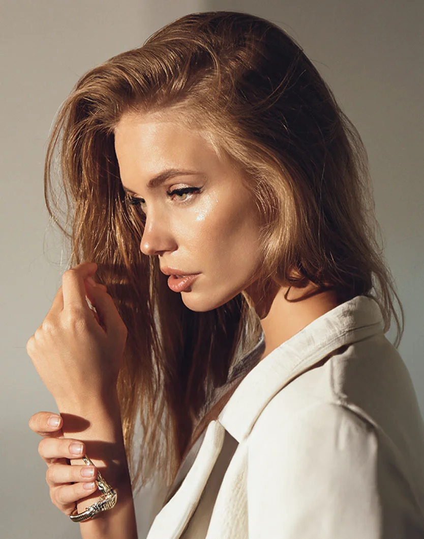 A light-skinned woman with long, voluminous light brown hair gazes off to the left with a neutral, yet inviting expression. She wears subtle makeup, including dark eyeliner, and a white blazer. A metallic bracelet is visible on her left wrist, with her hands gently placed near it. Soft, warm lighting highlights her face and hair, set against a light beige or gray background, giving the image a fashion portrait feel. - Radiesse in King of Russia, PA